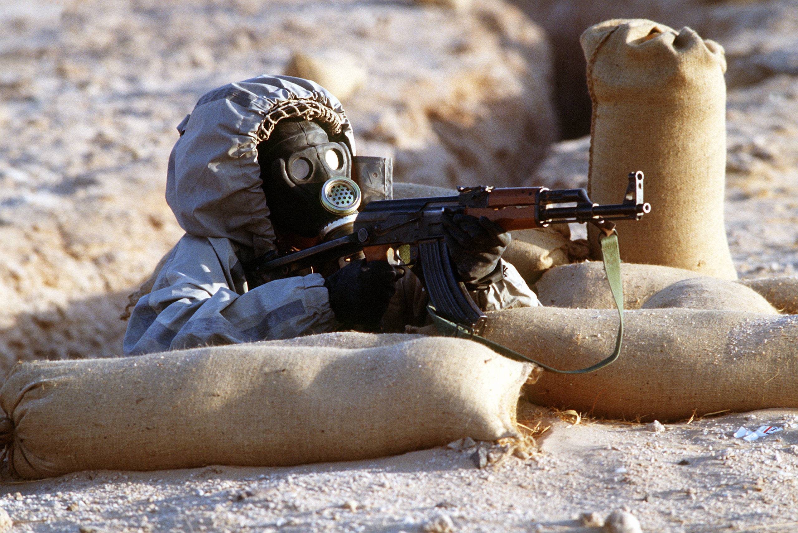 A Syrian soldier aims an AK-47 assault rifle from his position in a foxhole during a firepower demonstration, part of Operation Desert Shield.  The soldier is wearing a Soviet-made Model ShMS nuclear-biological-chemical warfare masks.