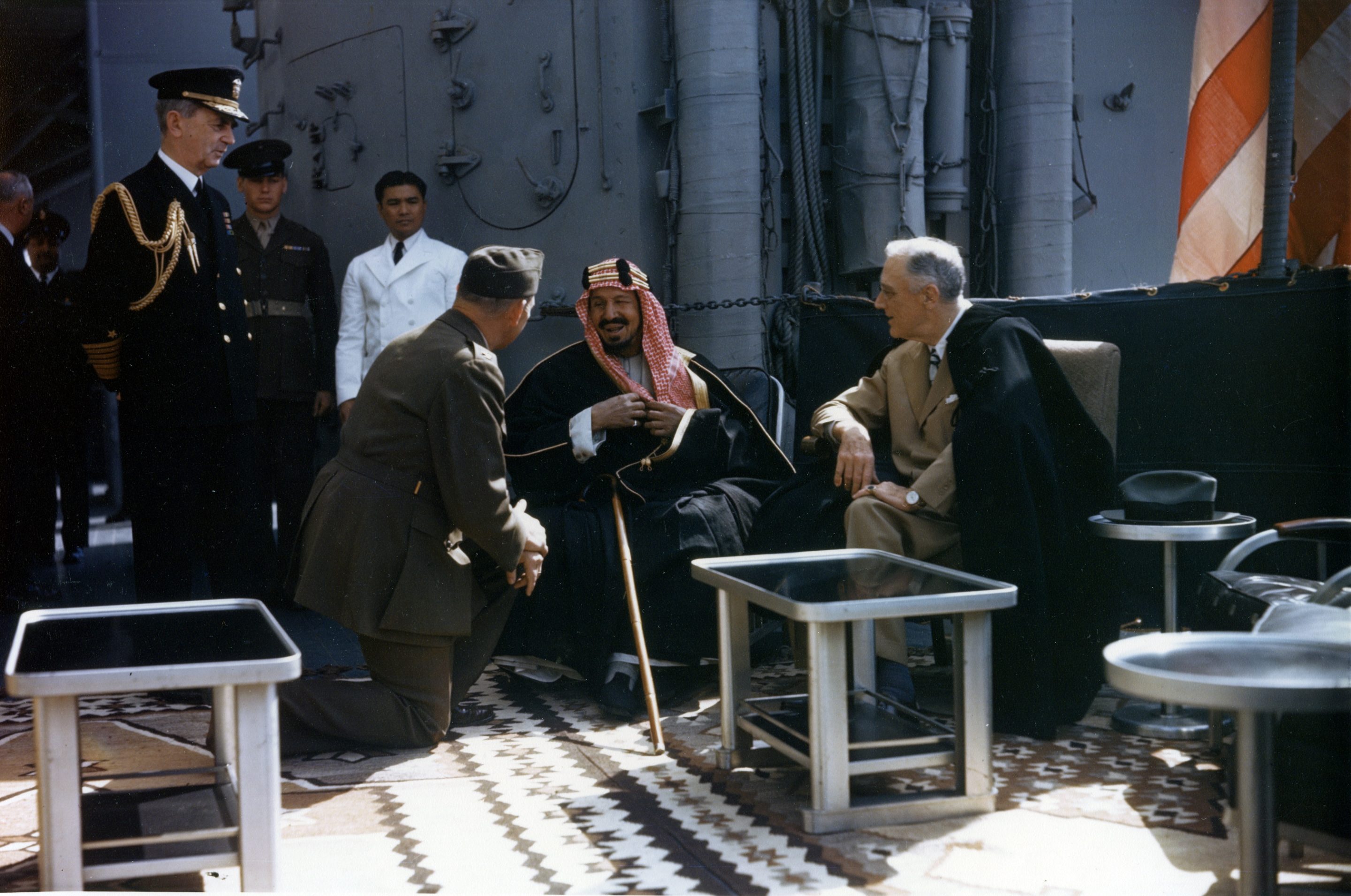 Le président Franklin Roosevelt en compagnie du roi Abdelaziz Al Saoud, du colonel William A. Eddy (en) (qui sert d'interprète, un genou au sol) et de l'amiral William Leahy (debout, à gauche) sur l'USS Quincy.